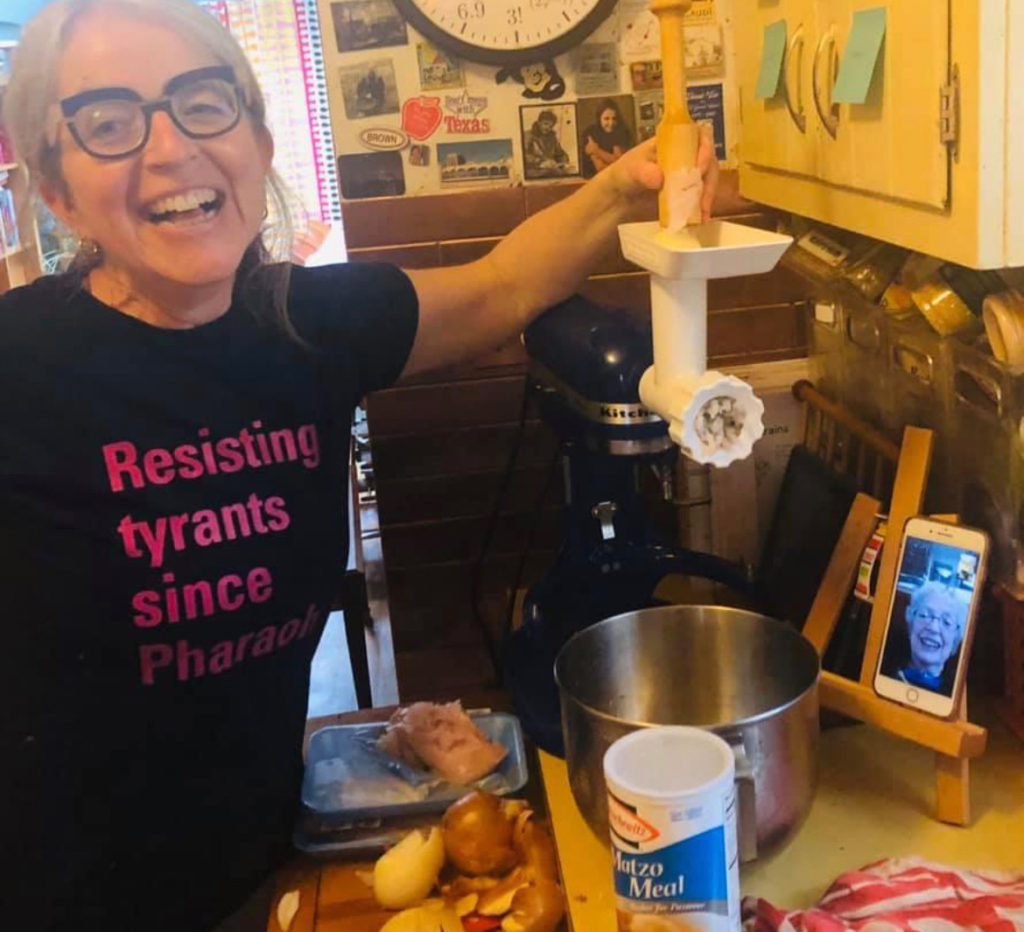 A middle aged white, Jewish woman preparing gefilte fish in a kitchen while being supervised by her mother over video chat. The woman is wearing a black t-shirt that has pink text that reads: Resisting tyrants since Pharaoh