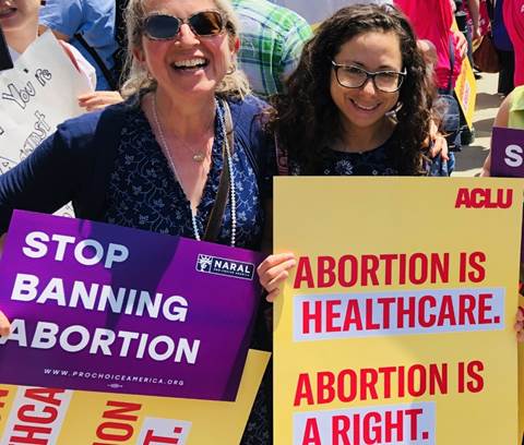 Two white women with pro-choice signs at a rally. Signs read "Stop Banning Abortion/ProChoiceAmerica.org / NARAL" and "Abortion is Healthcare / Abortion is a Right / ACLU."