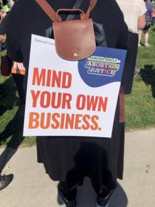 Photo of a woman's back, at a rally, with a large poster hanging from her backpack. Sign reads: "Jewish Rally for Abortion Justice / Mind Your Own Business." Washington, DC; May 2022