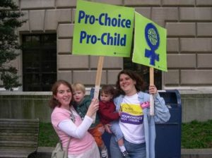 Color photo of a smiling, young white woman holding a small child and a Pro-Choice/Pro-Child sign while standing next to another smiling, young white woman holding a small child and a protest sign featuring the symbol for woman, a circle over a plus sign. 