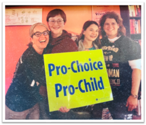 A group of four individuals, two white maternal figures on each end, and two young white teenagers in the middle, all standing together and smiling in front of a Pro-Choice/Pro-Child sign.