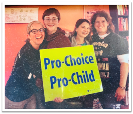 A group of four individuals, two white maternal figures on each end, and two young white teenagers in the middle, all standing together and smiling in front of a Pro-Choice/Pro-Child sign.