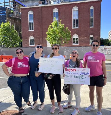 A group of four white women and one white man standing in a line, across the street from the Capital Jewish Museum's historic synagogue, and smiling at the camera. Two are holding pro-choice signs and two others are wearing pink "I stand with Planned Parenthood" t-shirts.