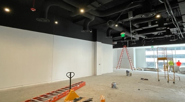 Interior view of a gallery space under construction. A ladder and miscellaneous equipment are scattered through out the space. The room has white walls and the ceiling features exposed systems and is painted black.