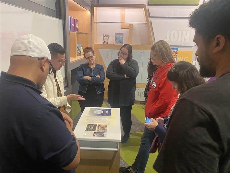 A curator stands on one side of a cart. The cart features documents and photographs. A group of six teachers look at the documents while the curator speaks about them.