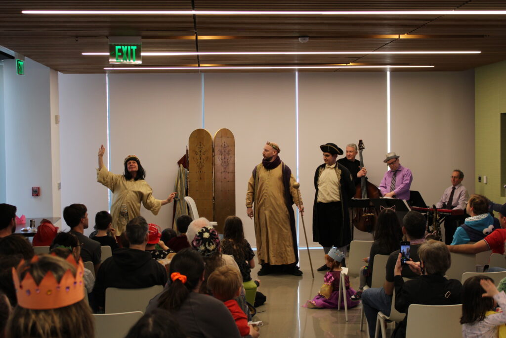 A group of performers in the background stand and act out a Purim spiel in robes and costumes. In the foreground sits an audience of children and grown ups.