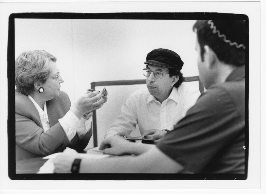 A black and white photograph of a women and two men sitting a a table discussing together. The woman is to the left with one man in the center and the other with his back to the camera to the right.
