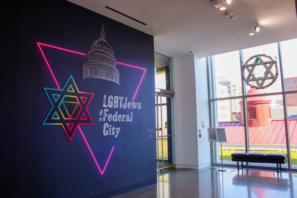 Photo of the LGBTJews in the Federal City exhibition entrance and title wall, featuring large illustration using the exhibition’s wordmark, an illustration of a neon pink triangle, a line drawing of the Capitol building, and a colorful Jewish star with a triangle at its center.