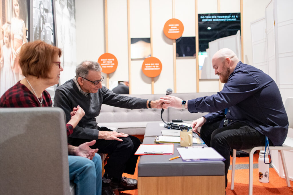 In a museum gallery, a man, sitting next to a woman on a sofa, hands a microphone to a man sitting across from him. Papers and files sit on the table between them.