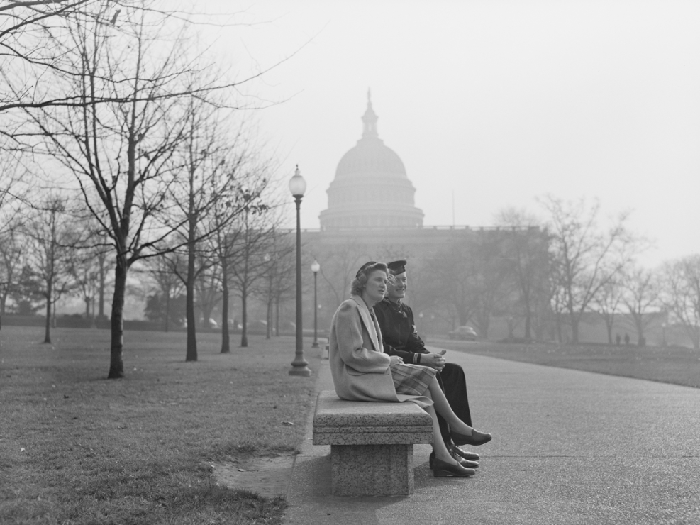 Black and White photograph of a man in a military uniform and a woman dressed in 1940's attire sitting on a stone bench in front of the US Capitol building.