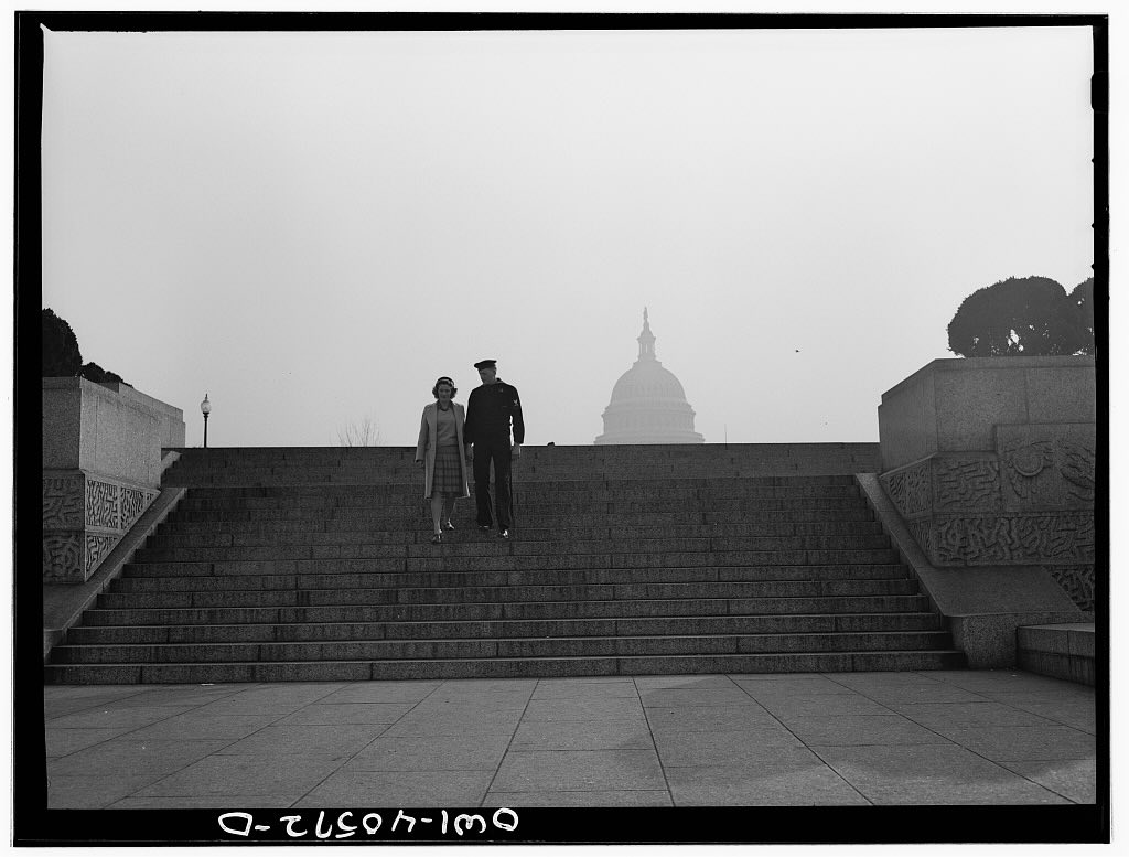 Black and white photo of a man and a woman descending a wide staircase with a view of the U.S. Capitol in the background. The man is wearing a soldier's uniform.