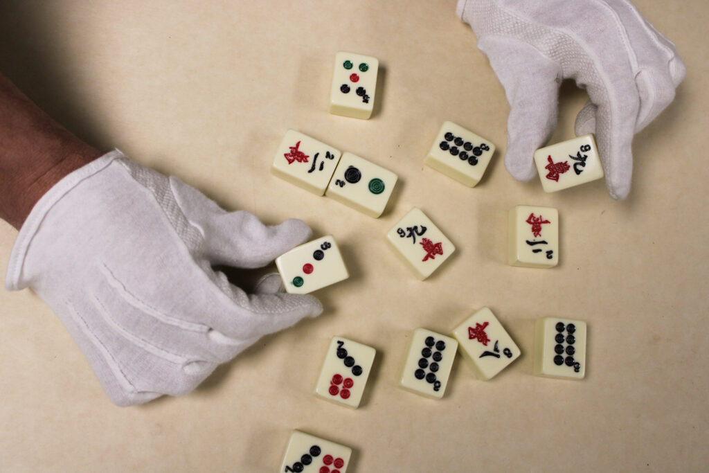 A collection of mahjong tiles on a beige surface. Two hands in white cloth gloves hold two tiles.